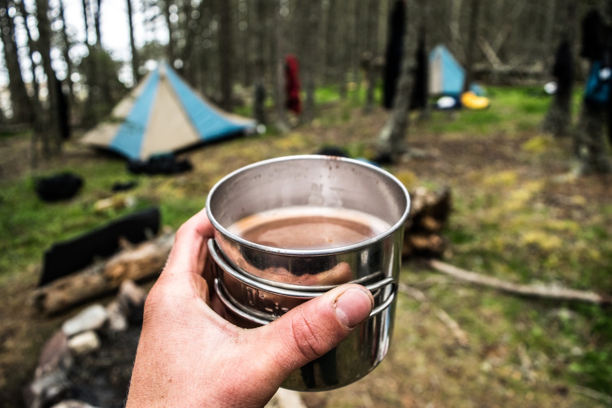 hand holding tin cup of hot coffee with blue and tan mega mid tent in the background