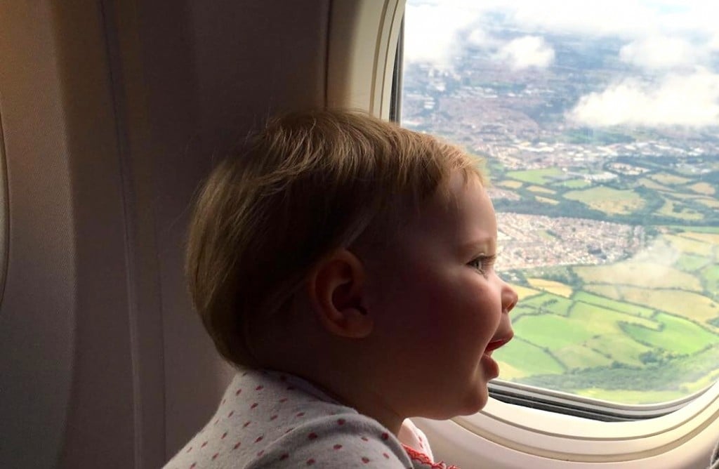 Baby smiles and looks out an airplane window at patchwork of green fields