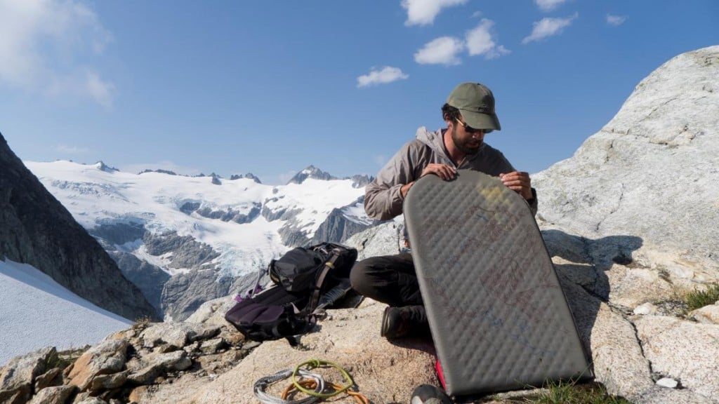 NOLS participant sits on rocky outcrop holding inflatable sleeping pad with snowy mountains behind