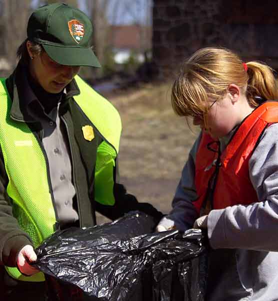 Park Service ranger and child wearing red vest and glasses hold trash bag together