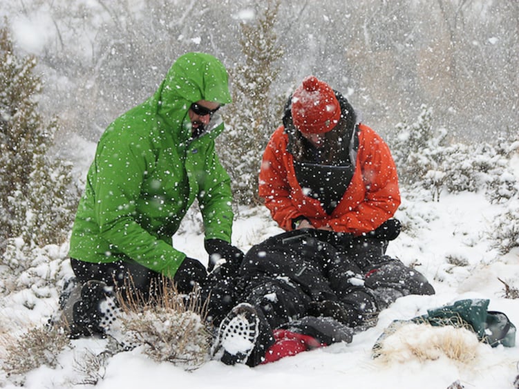 Two rescuers kneel in a snowy forrest while treating a collapsed patient.