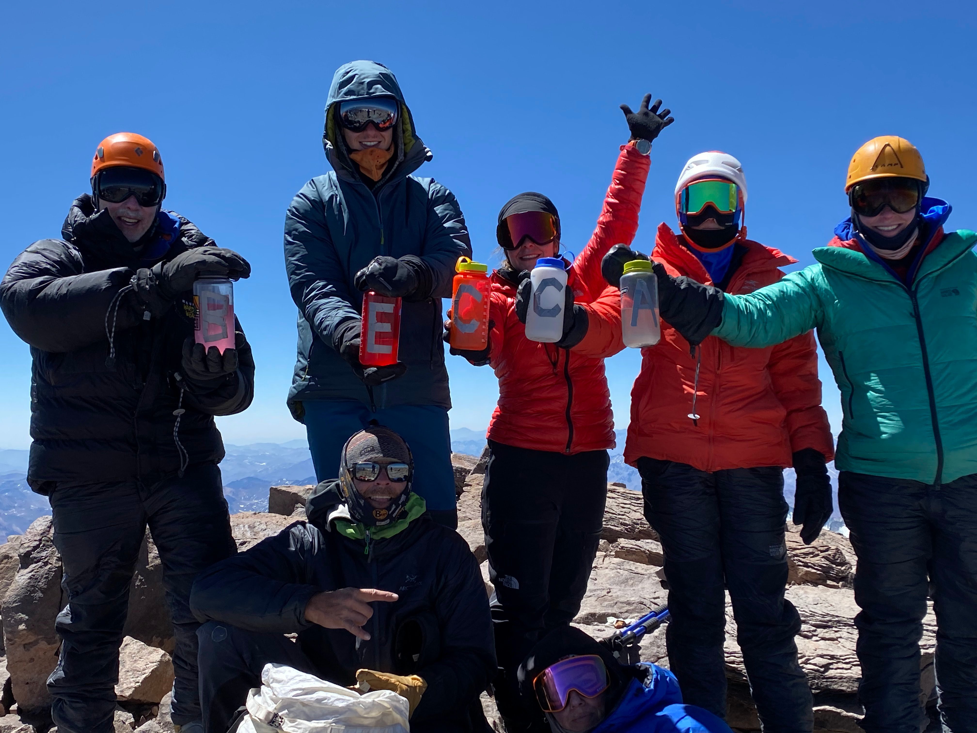 Six people standing at the summit of Aconcagua holding up water bottles with letters to spell: BECCA