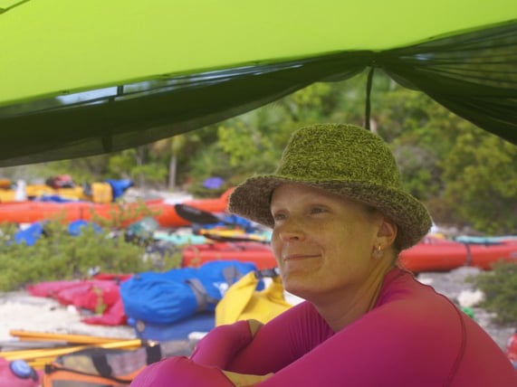 Woman in a beach hat sitting under a tarp smiling as she looks out the ocean