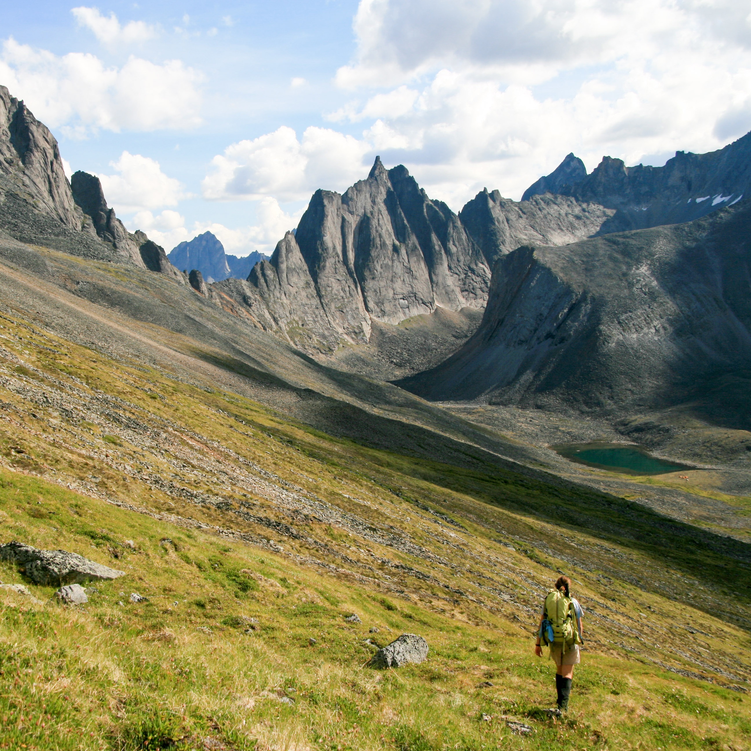 Backpacker hikes through grassy foothills in the Yukon wiht high, steep peaks in the background