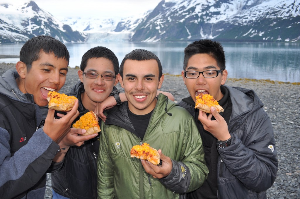 Four teens eat slices of pizza on a lakeshore
