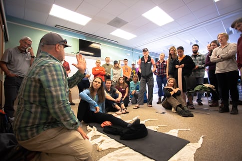A group of students stand around a person who is lying on the floor. A man in the foreground is using his hands to show what a spine looks like
