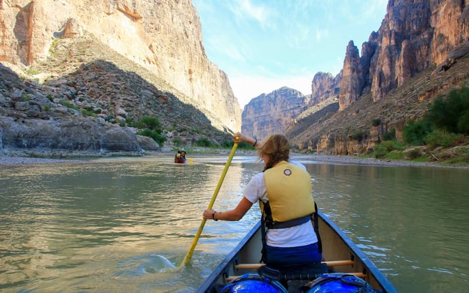 A canoeist paddles down a tan river between tall cliffs
