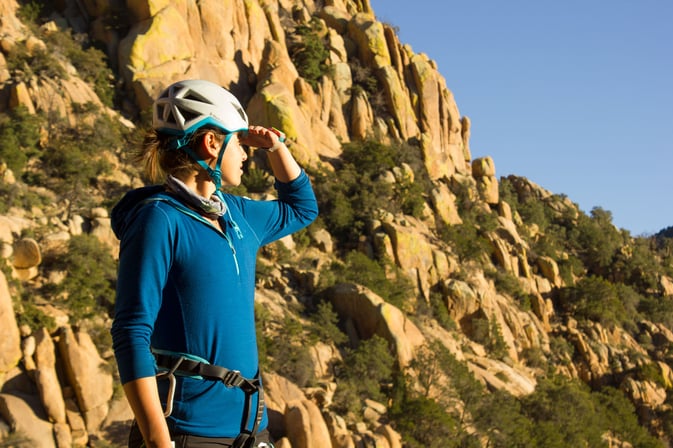 a person wearing a climbing harness and a helmet stands in front of mountains and looks out over the surrounding area