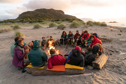 A group sits in the sand around a fire. There is a sand dune in the background and the sun is setting