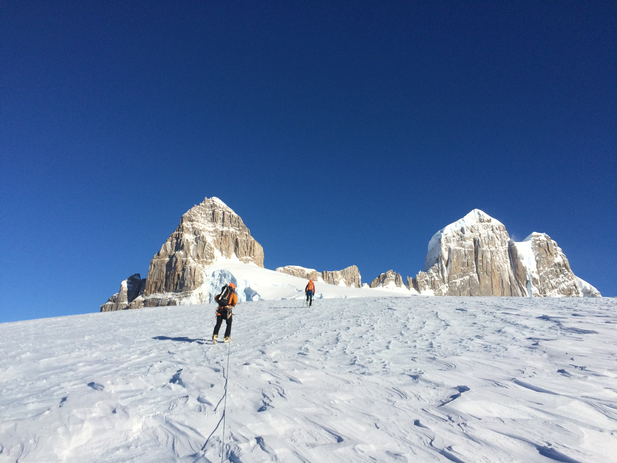 two climbers on a rope team ascend a steep snowfield toward rocky peak in Patagonia