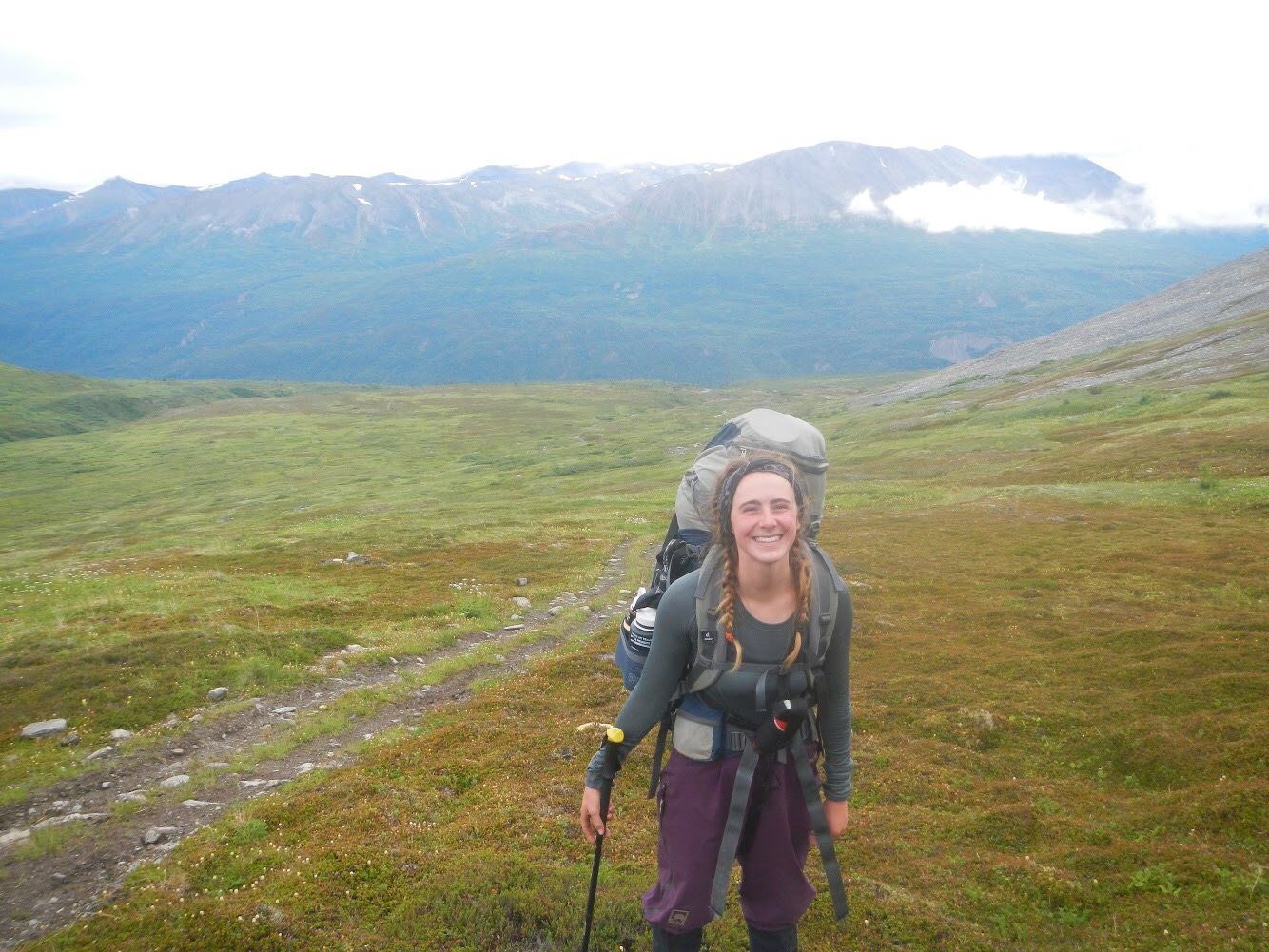 Woman with hair in braids wearing a backpack and smiling next to a trail in Alaska's mountains