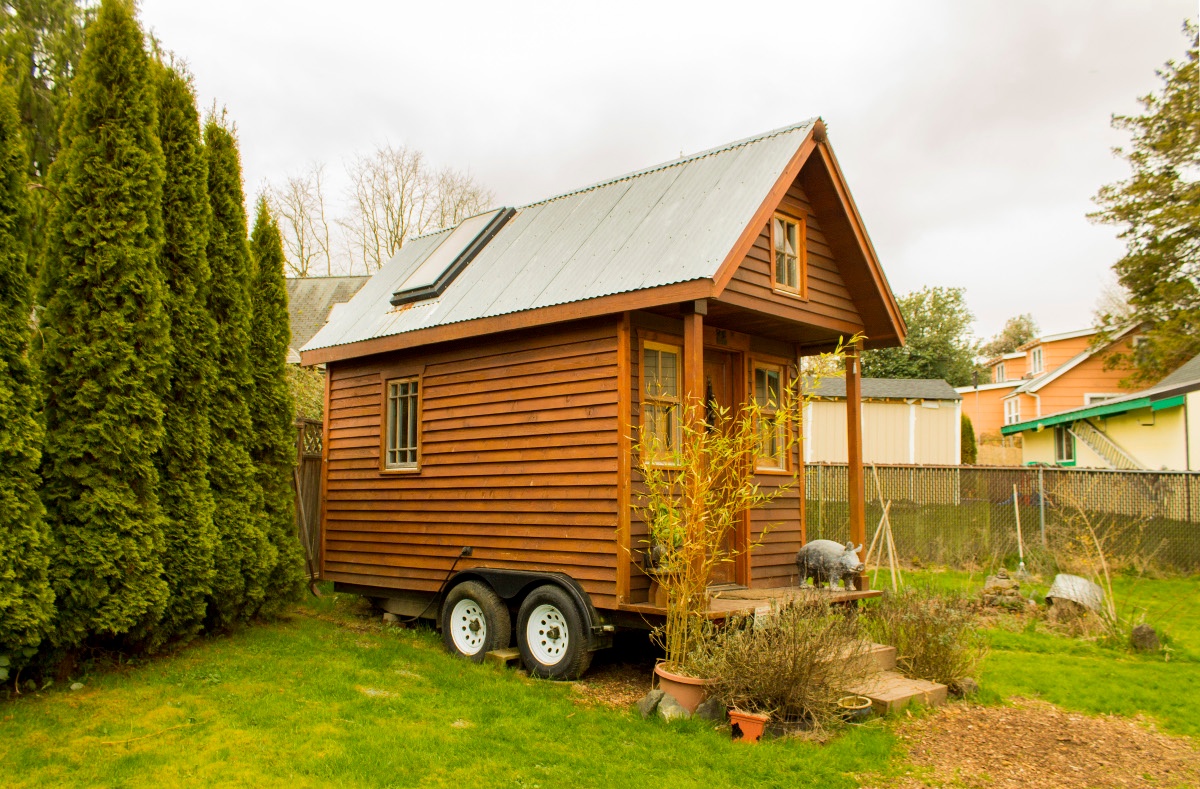 tiny house with porch parked on a trailer on green grass near a hedge