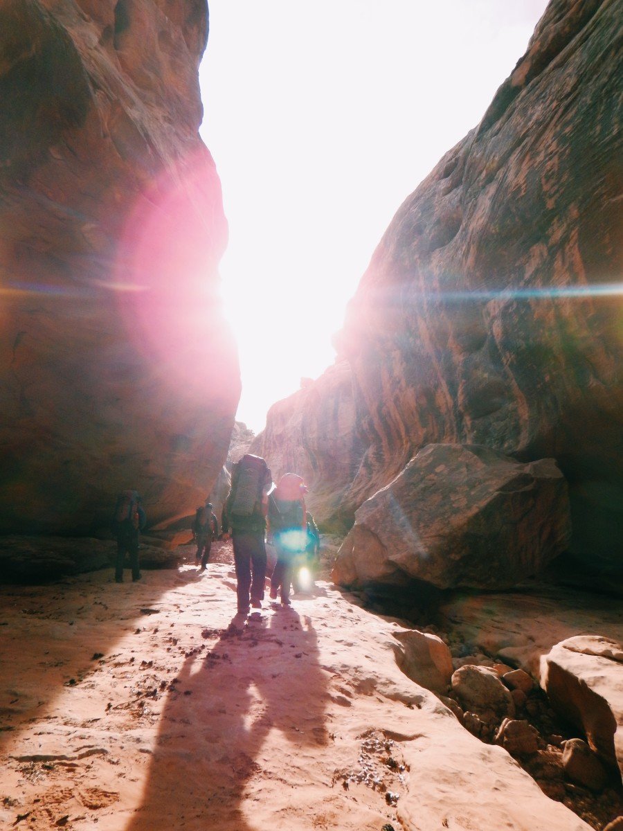 Students hike through a red rock canyon, the sun at their faces with long shadows trailing behind