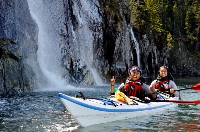 two smiling NOLS students in a white double sea kayak next to a waterfall in Alaska