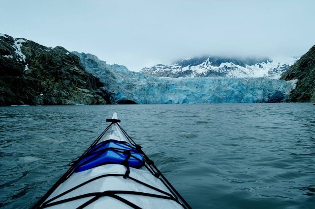 looking over the nose of a sea kayak toward blue glacier and mountains partly obscured by fog