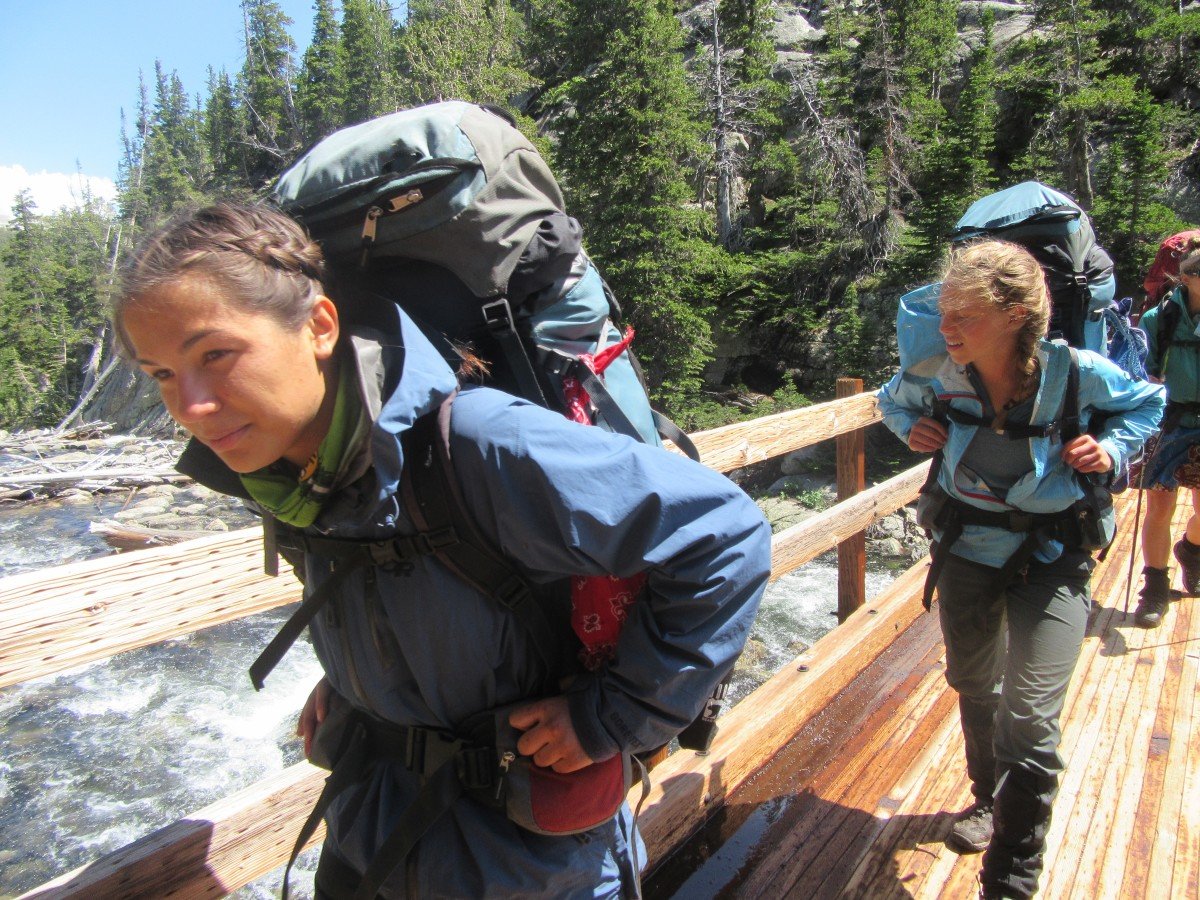 two teenage girls with backpacks walk on a wooden bridge over a river