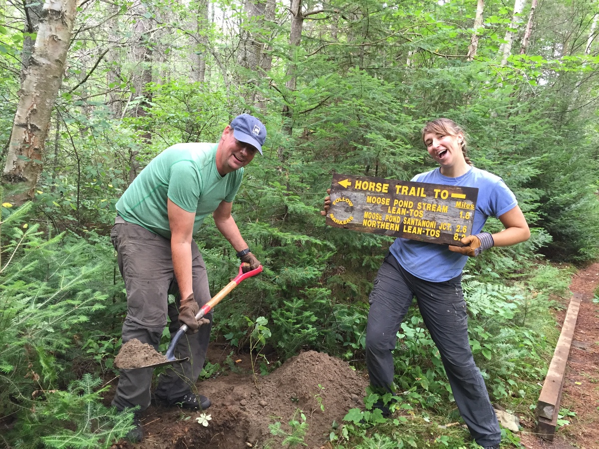 two smiling people work on a service project with man using a shovel and woman holding a trail sign