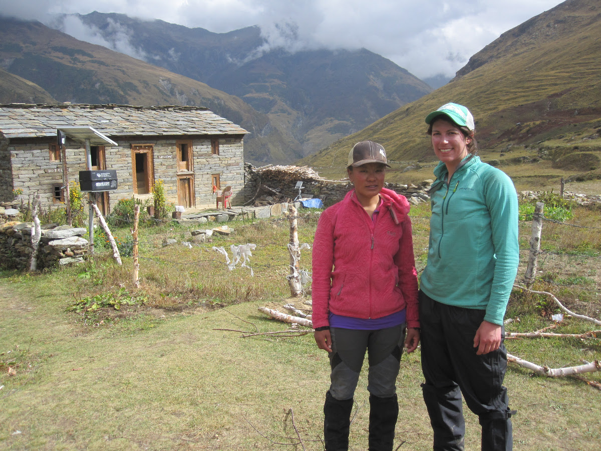 two women stand together near a stone and wood house in the Himalayas