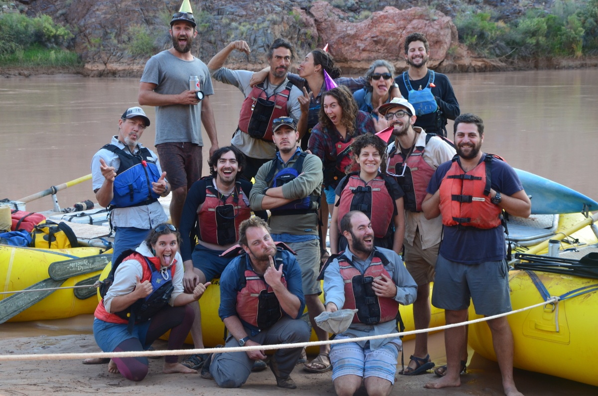 Group of people making goofy faces and wearing life preservers next to rafts on a river trip