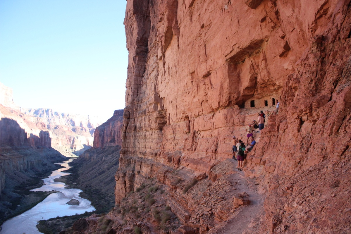 Hikers follow a narrow path winding along a rocky red canyon wall in the Grand Canyon