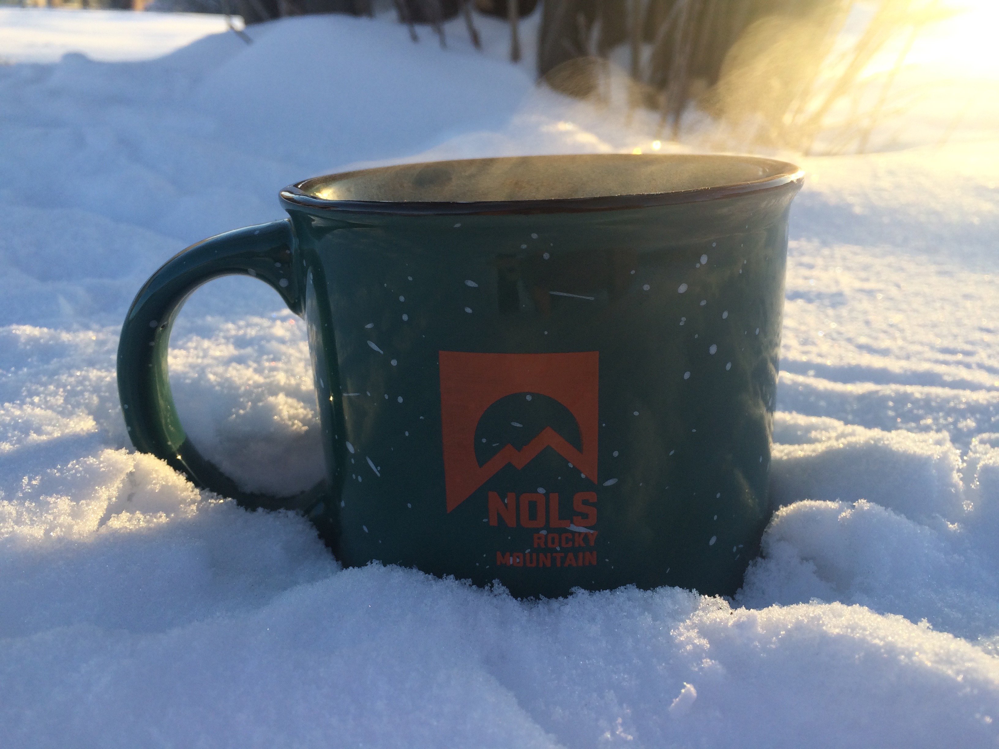 green coffee mug with orange NOLS logo sitting in the snow with steam rising off it