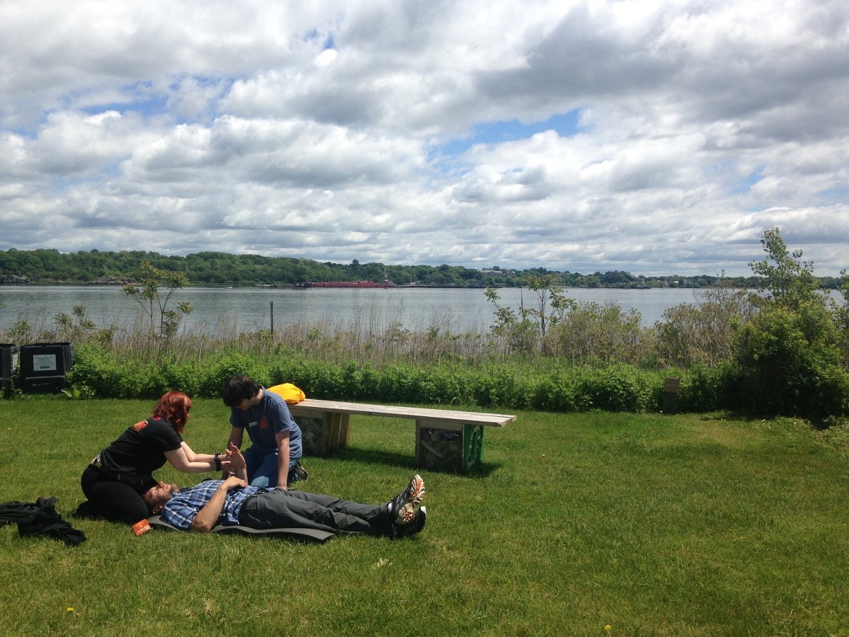 two NOLS Wilderness Medicine students practice caring for a mock patient lying in the grass with the Narragansett Bay behind