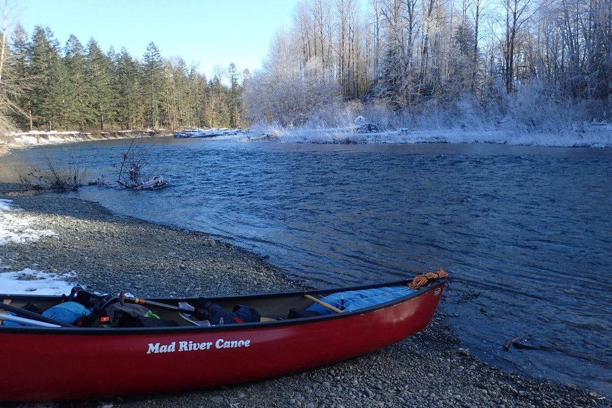 Red Mad River canoe pulled up on a pebbly shore along a river lined with pines