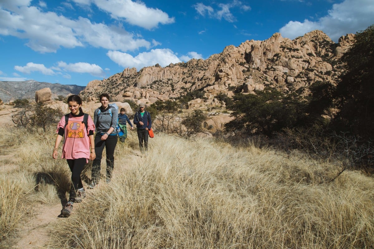 Students hike along a single track trail past dry grass and reddish rock outcroppings in the Southwest