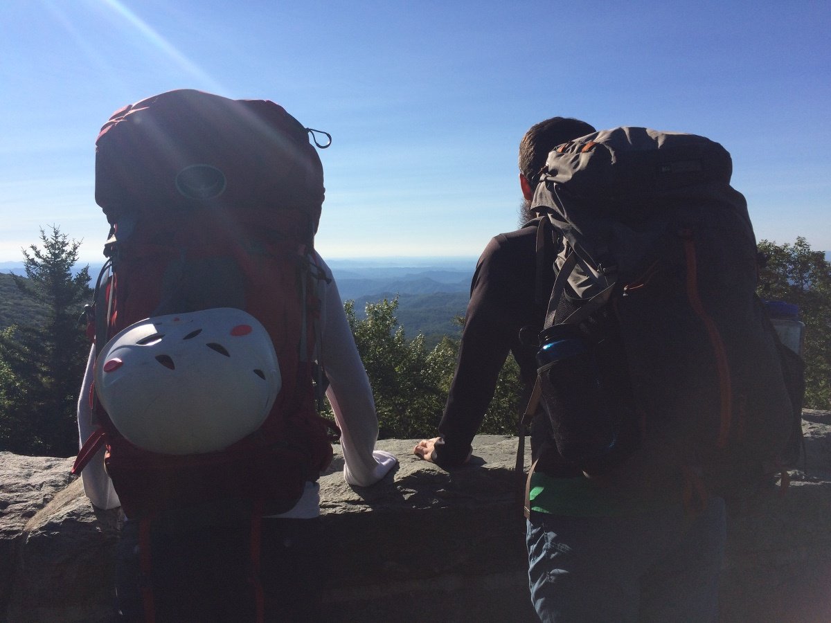 two people with backpacks stand with hands on a low wall in front of them, looking out at distant mountains