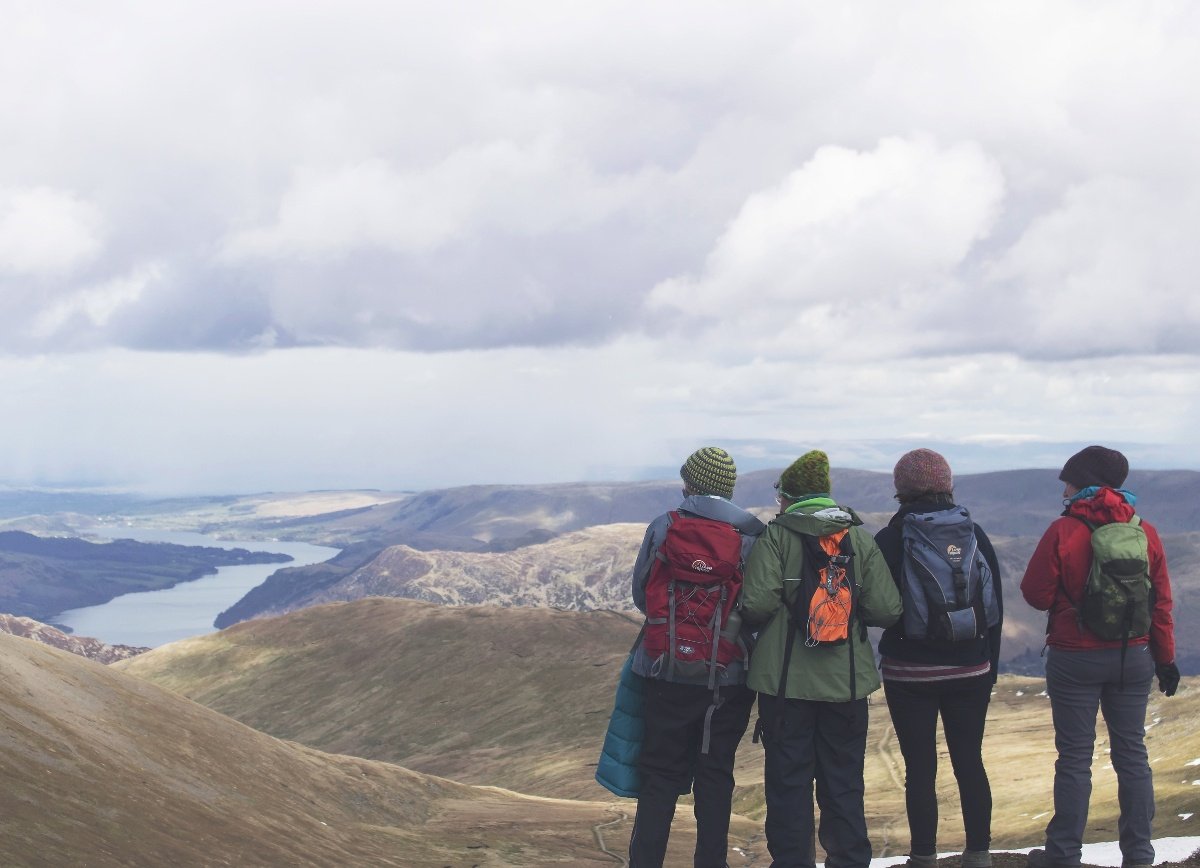 four students carrying backpacks stand with their backs to the camera looking out over barren hills to water below