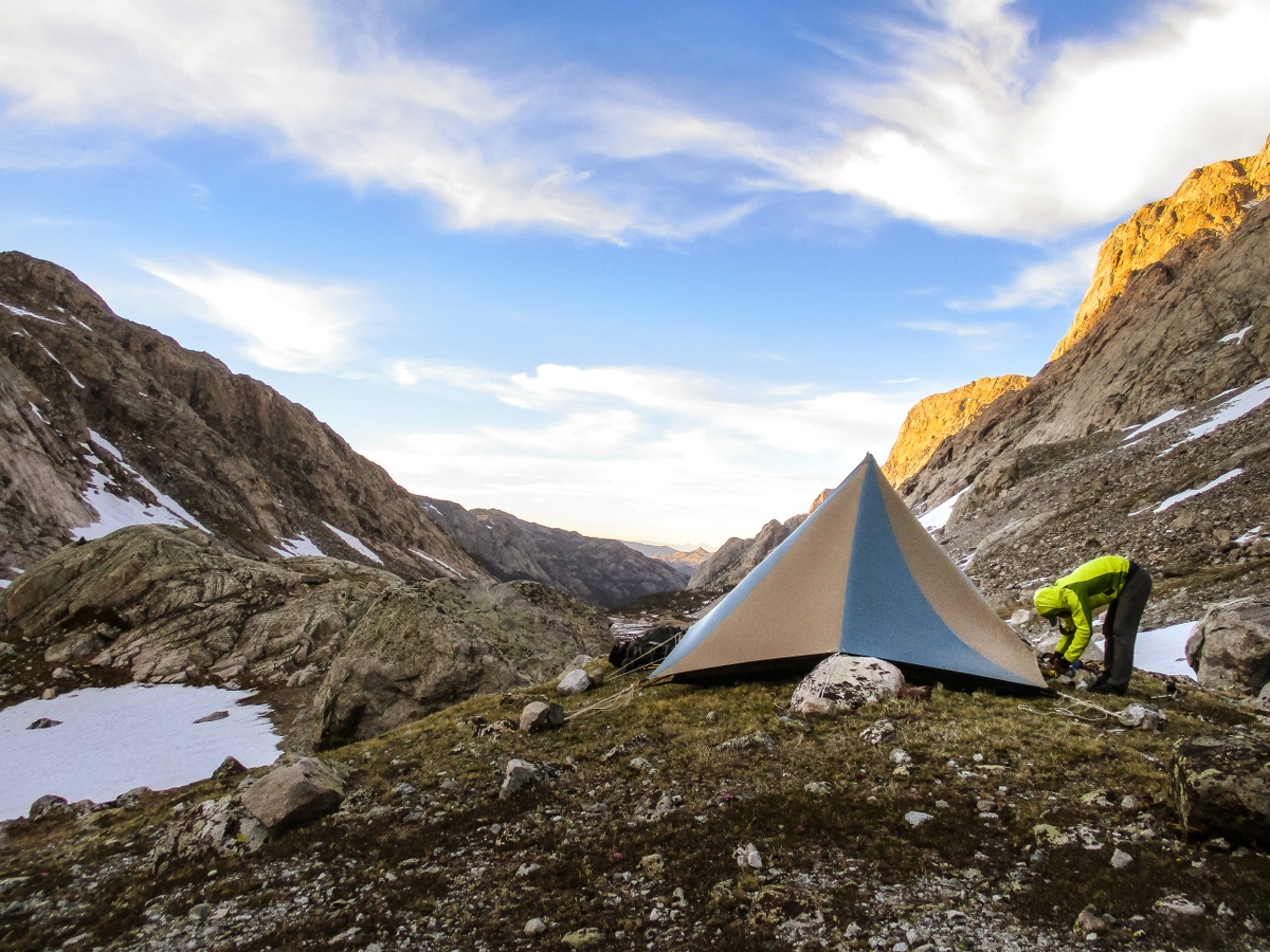 NOLS student wearing neon jacket secures blue and tan mega-mid tent in a rocky camp in the mountains