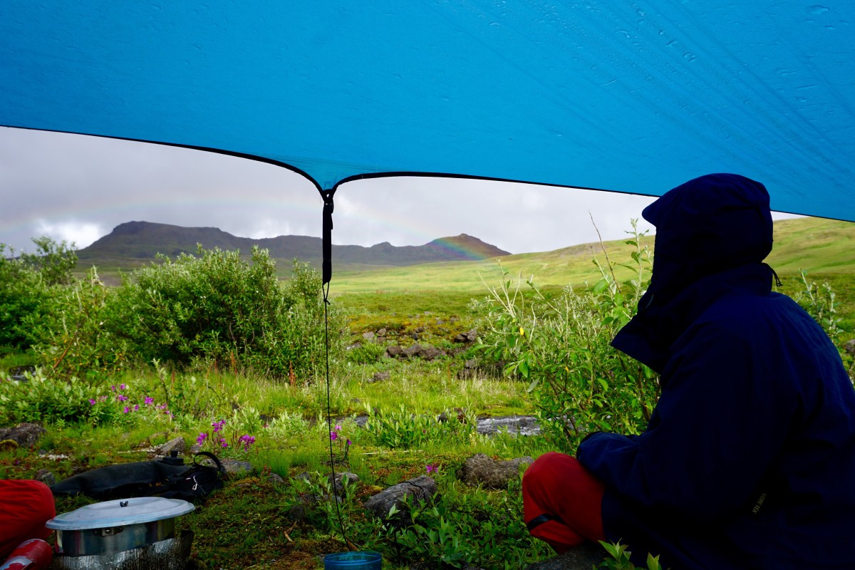 hunkering under a tarp next to cooking supplies while looking out at green vegetation and a rainbow in Alaska
