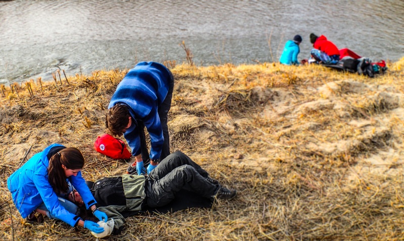 two small groups of NOLS wilderness medicine students practice caring for mock patients on a grassy shore beside a river