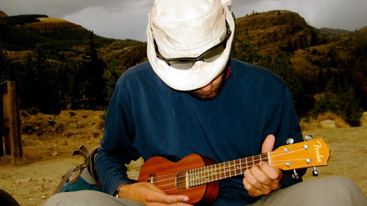 Man wearing hat with sunglasses on top strums a ukulele outdoors