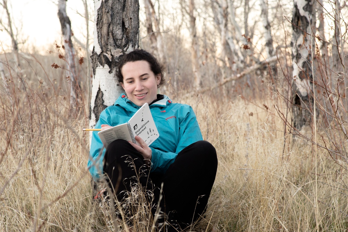 smiling student wearing a turquoise jacket leans against an aspen tree and writes in a journal