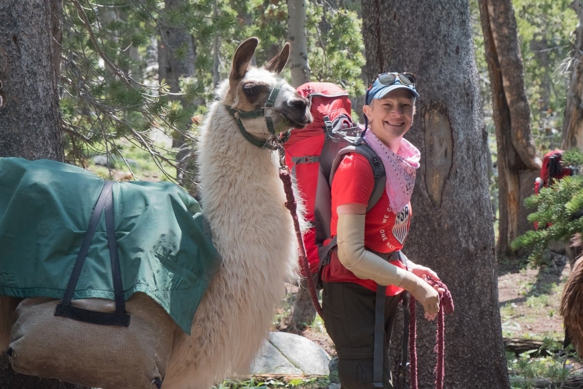 smiling woman with backpack, pink bandana, and baseball cap stands next to pack llama