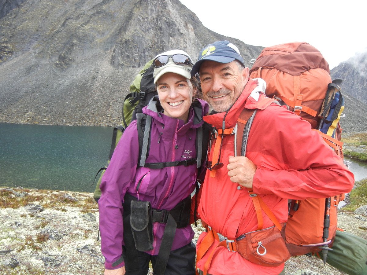 Two people smile wearing backpacks and rain jackets in front of a mountain lake