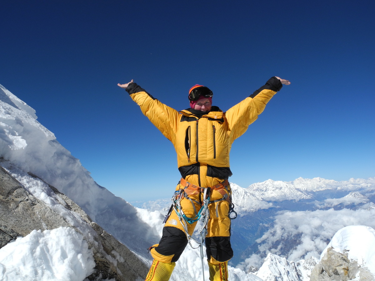 Woman wearing yellow snow gear smiles and raises her arms in the air on a sunny day high in the mountains