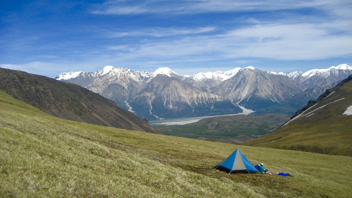 Blue and tan mega mid tent set up on a grassy slope in Alaska with snow-capped peaks beyond