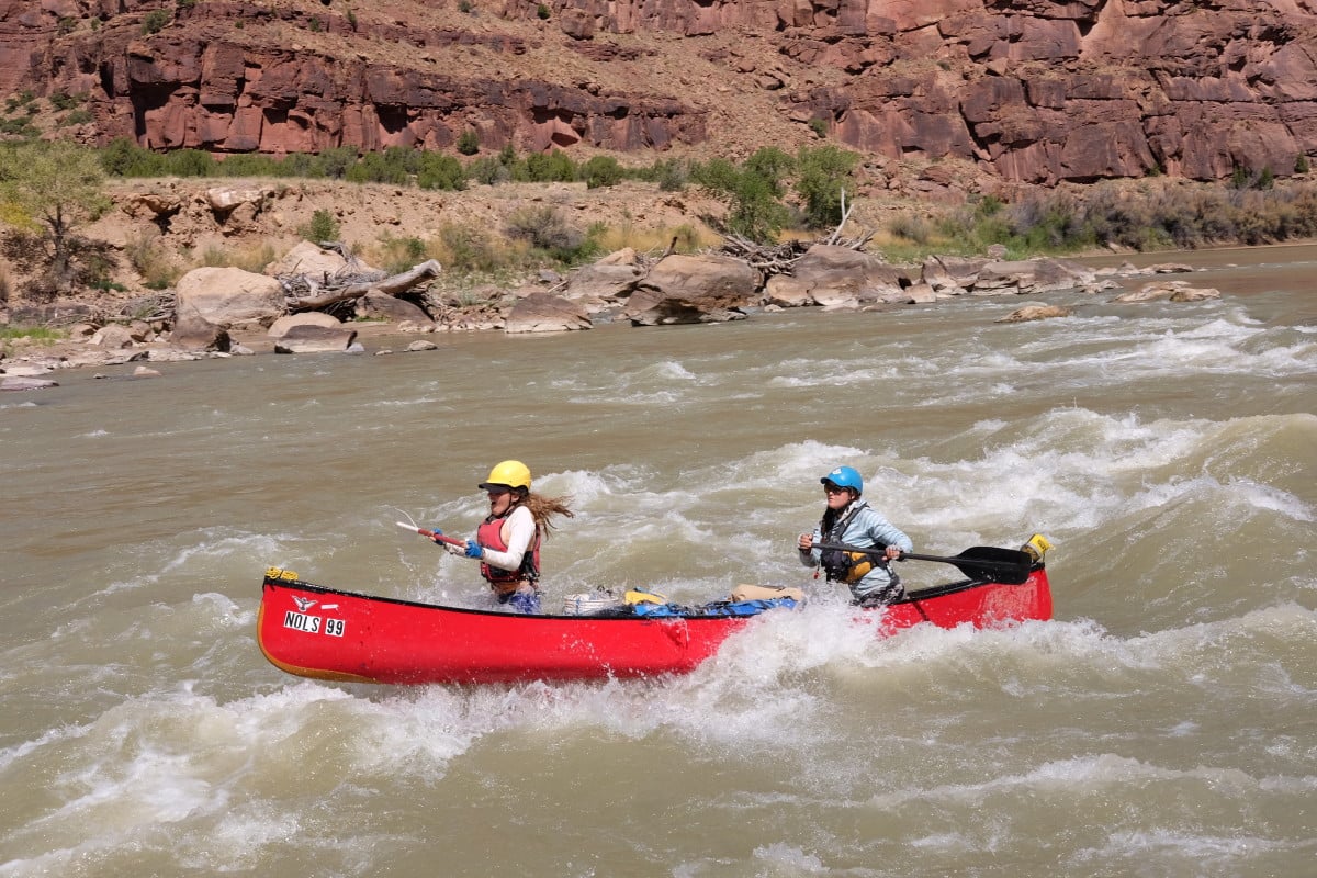 NOLS student and instructor paddle a red canoe through a rapid in a river canyon