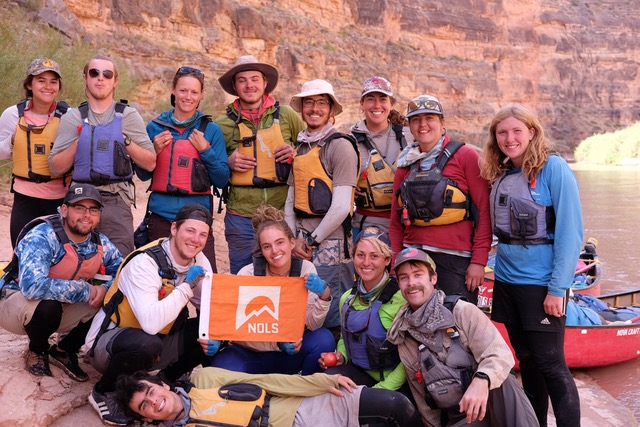 Author's group smiles for a group shot holding an orange NOLS flag while canoeing the Green River