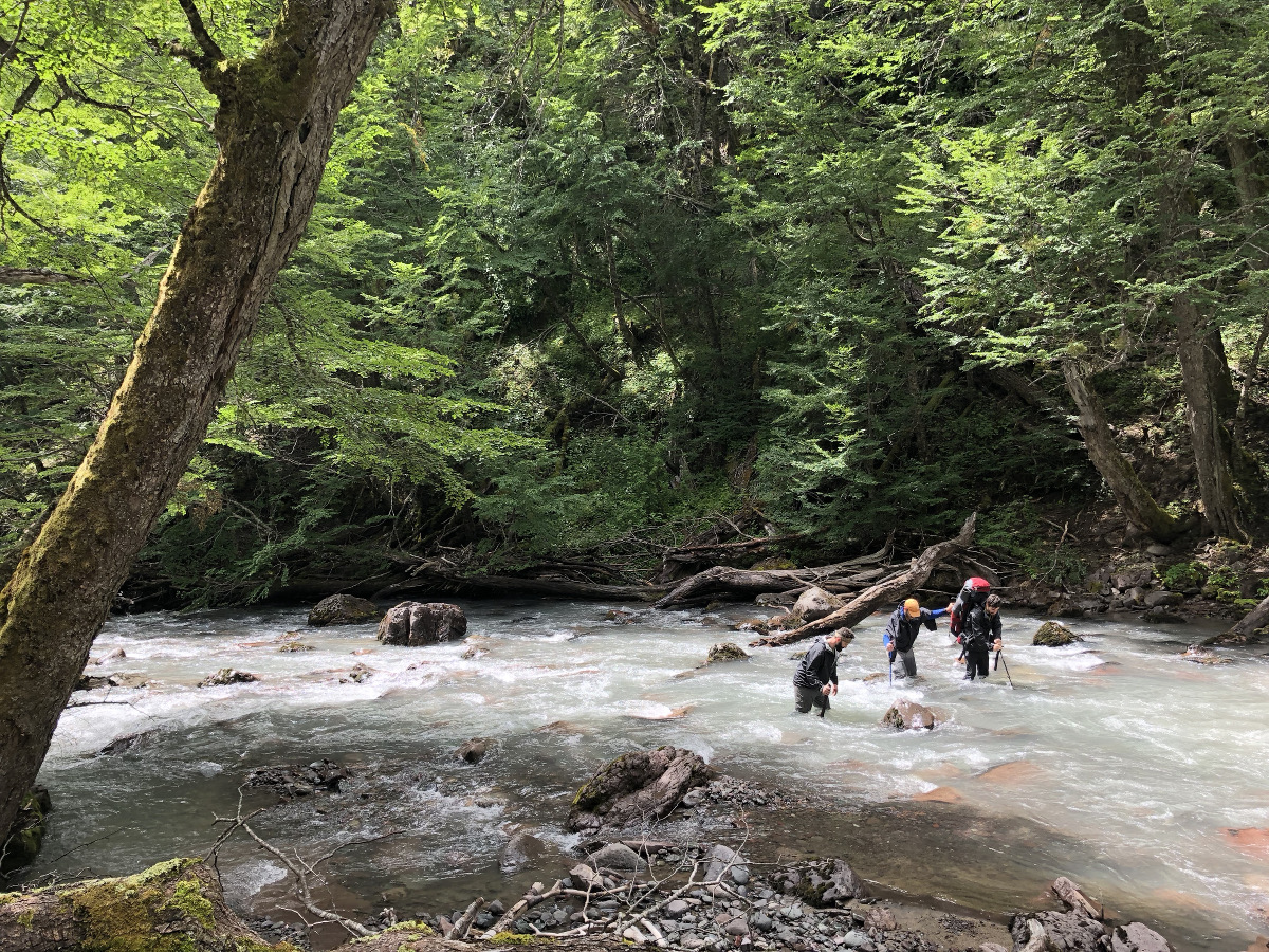 three students work together and use trekking poles to cross a creek in the mountains