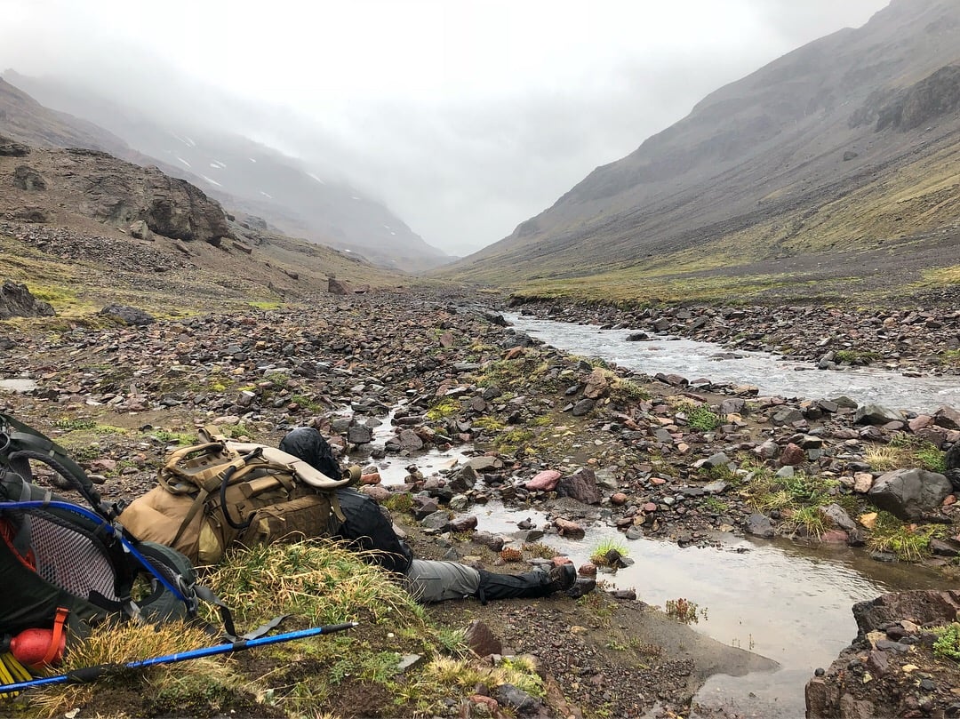 Student leans up against his backpack for a rest near a rocky stream on a foggy day in Patagonia