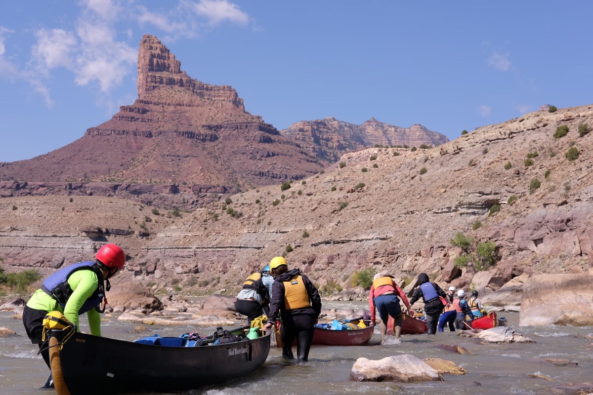 A group of people walks canoes through a rapid on a river in the desert