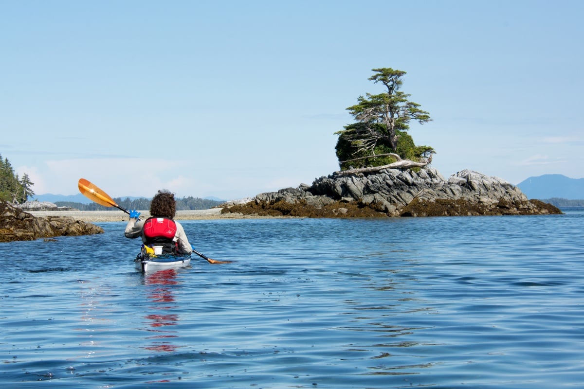 Paddler kayaks on calm blue water toward a rocky island with bonsai tree