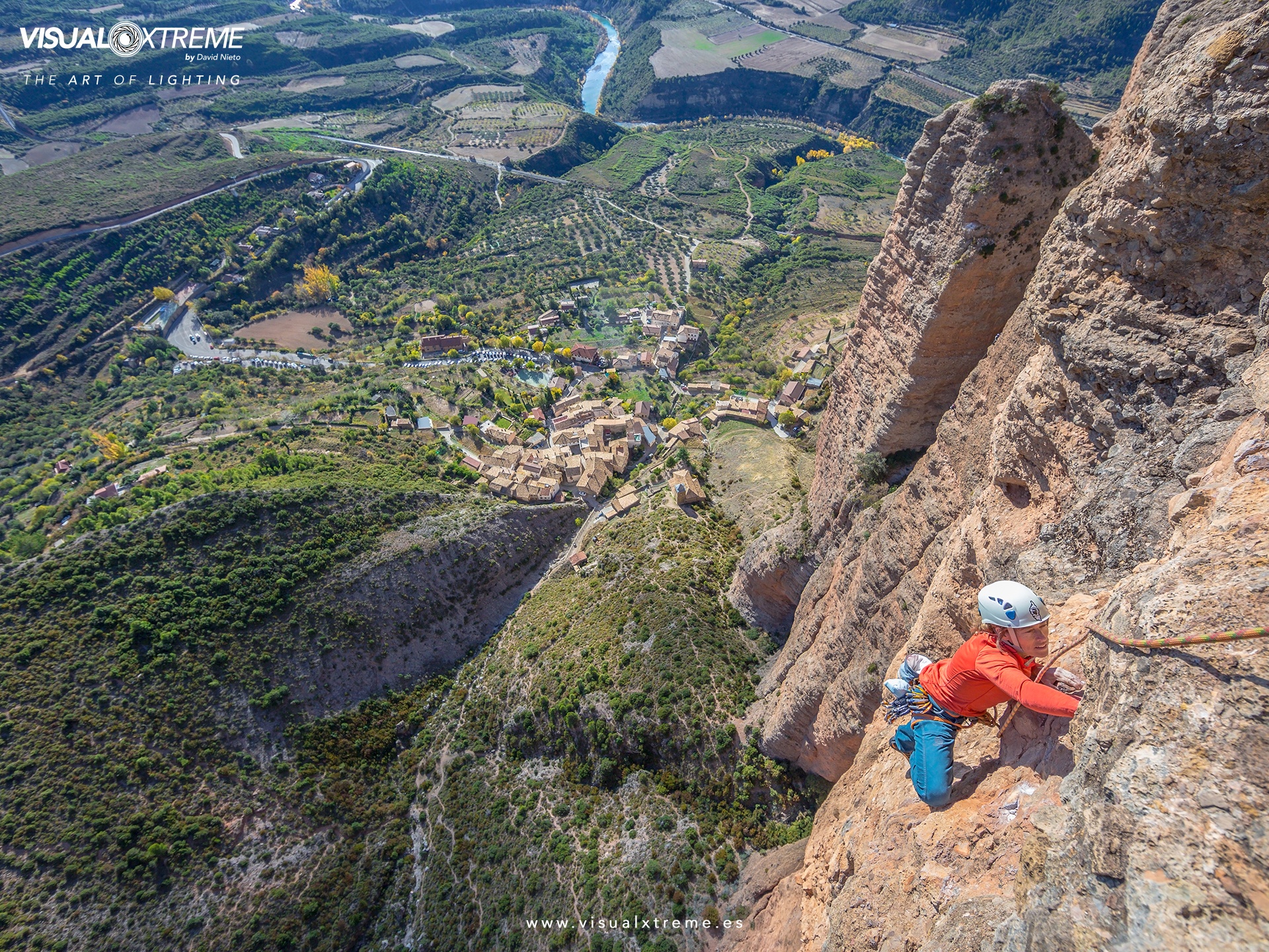 woman rock climbs on reddish rock face high above a small town