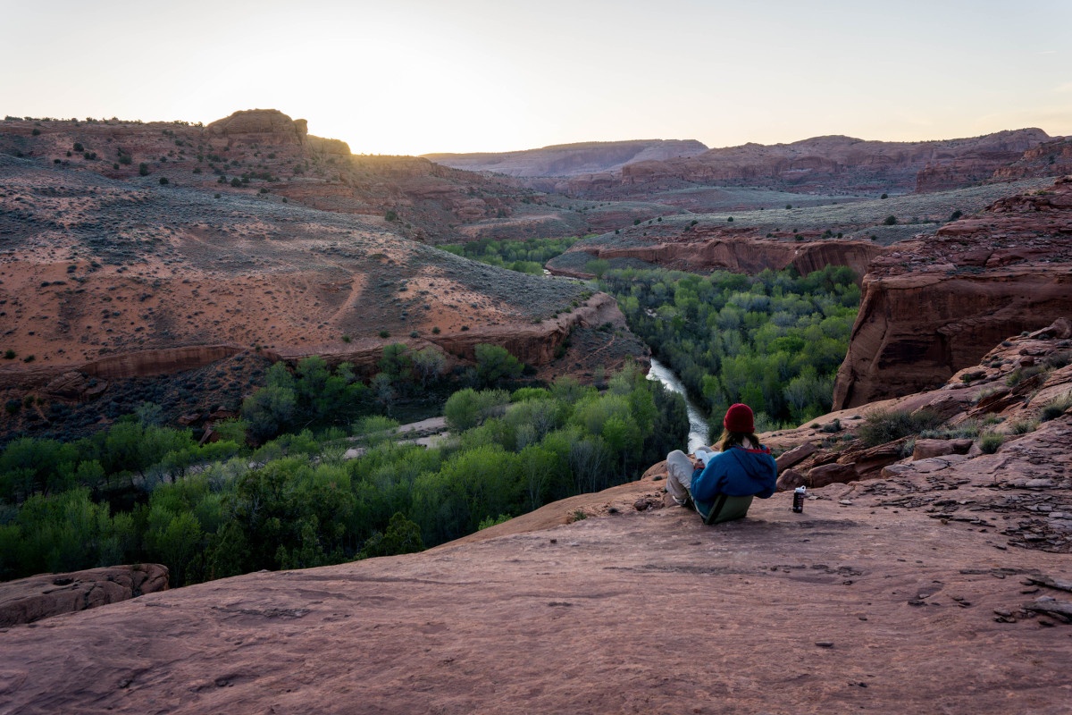 NOLS student sits on flat red rock formation looking out at green trees lining a red rock canyon and writes in her journal