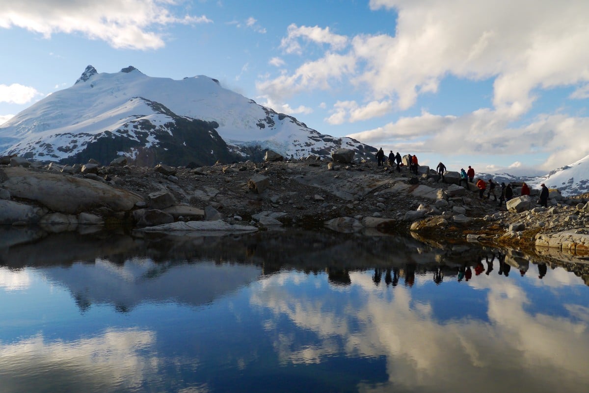 distant figures hike up a rocky slope toward snowy mountains surrounding a glassy lake with clouds reflected on its surface