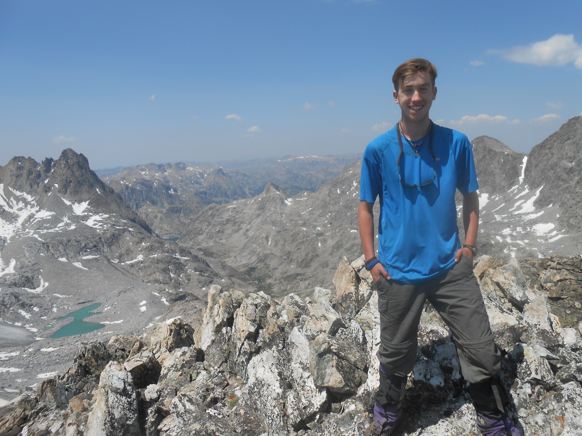 smiling student stands on a rocky outcrop with a turquoise alpine lake behind him in Wyoming's Wind River Range