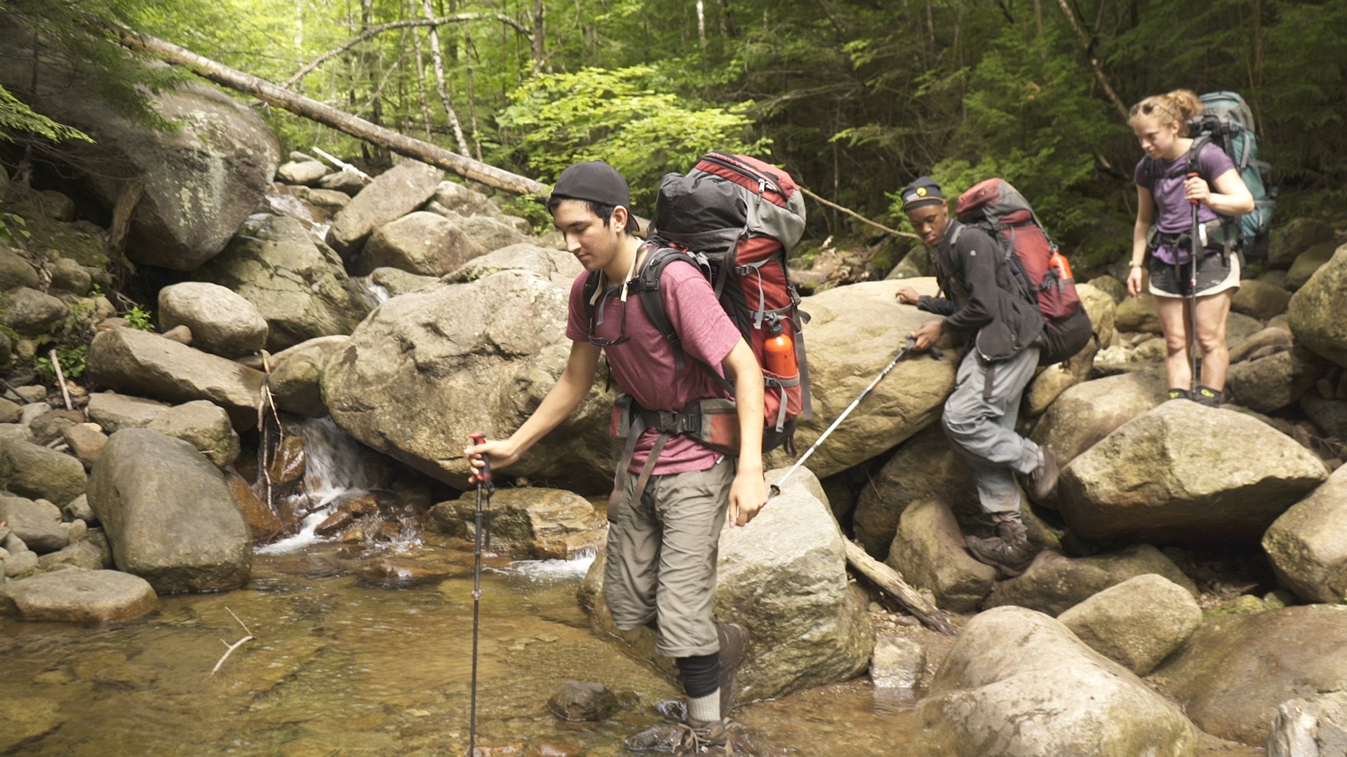 students wearing backpacks use trekking poles to cross a rocky creek in the Adirondacks